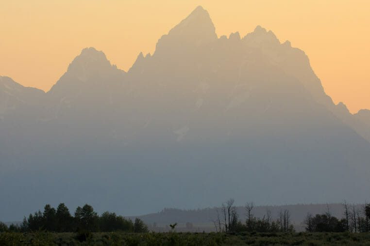 The Iconic Peaks Of The Teton Range Are Silhouetted Against A Warmly Lit Sunset In Grand Teton National Park